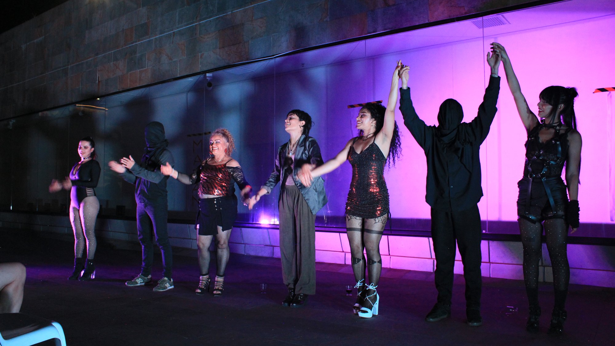 Grupo de artistas saluda con las manos unidas sobre un escenario iluminado de violeta.