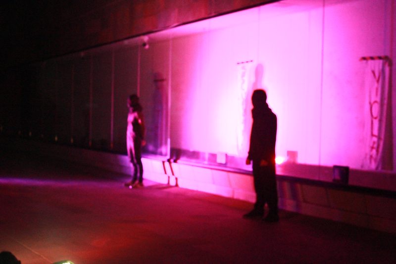 Dos figuras se encuentran frente a una pared iluminada de rosa.