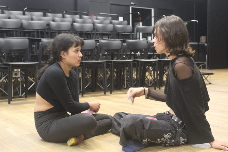 Dos personas adultas conversan sentadas sobre un escenario.