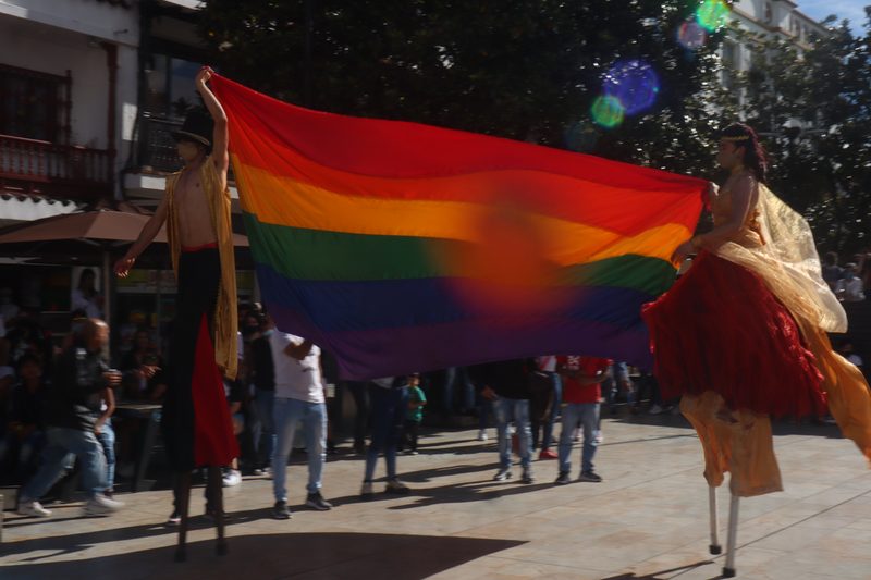 Dos artistas sobre zancos sostienen una bandera multicolor en una plaza.